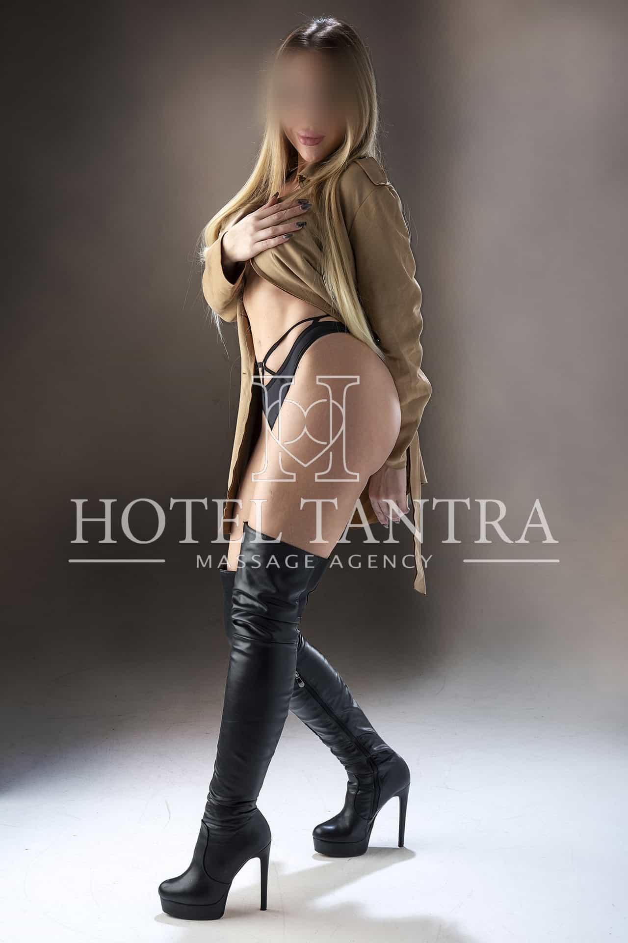 Blonde model with blurred face wearing a tan trench coat over black lingerie and thigh-high boots, posing sideways in a studio.