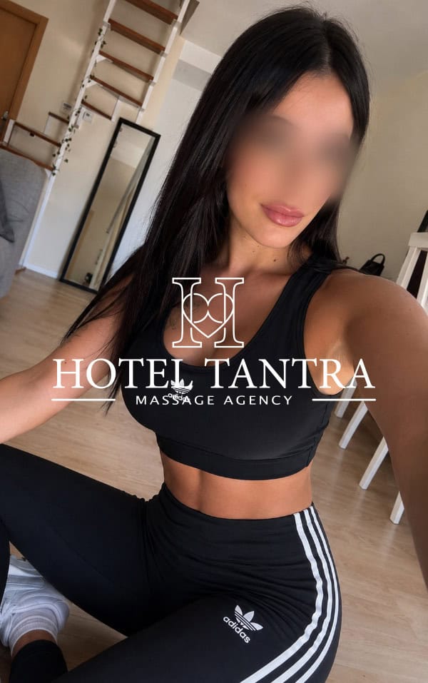 Outcall-massage-by-Lola 1 - Erotic Massage & Outcall Massage in Madrid Woman with long dark hair in a black sports bra and black Adidas leggings sits on the floor indoors; face blurred, watermark reads Hotel Tantra Massage Agency.