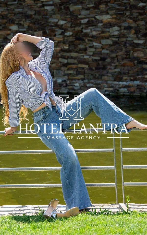 Footjob-madrid-anastasia 1 - Erotic Massage & Outcall Massage in Madrid Woman in a striped shirt and blue jeans posing on a railing beside a greenish body of water, with the words 'Hotel Tantra Massage Agency' as a logo overlay.