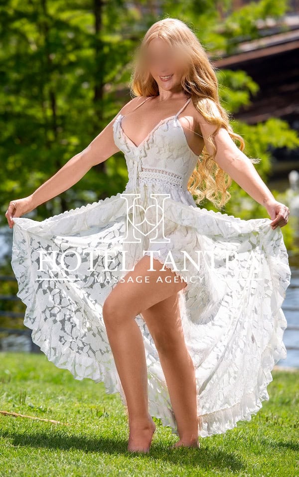 Hotel-tantra-anastasia - Erotic Massage & Outcall Massage in Madrid Woman in a white lace dress stands on grass outdoors, lifting the skirt to reveal its flowy length. A watermark across the center reads 'HOTEL TANNER MASSAGE AGENCY'.