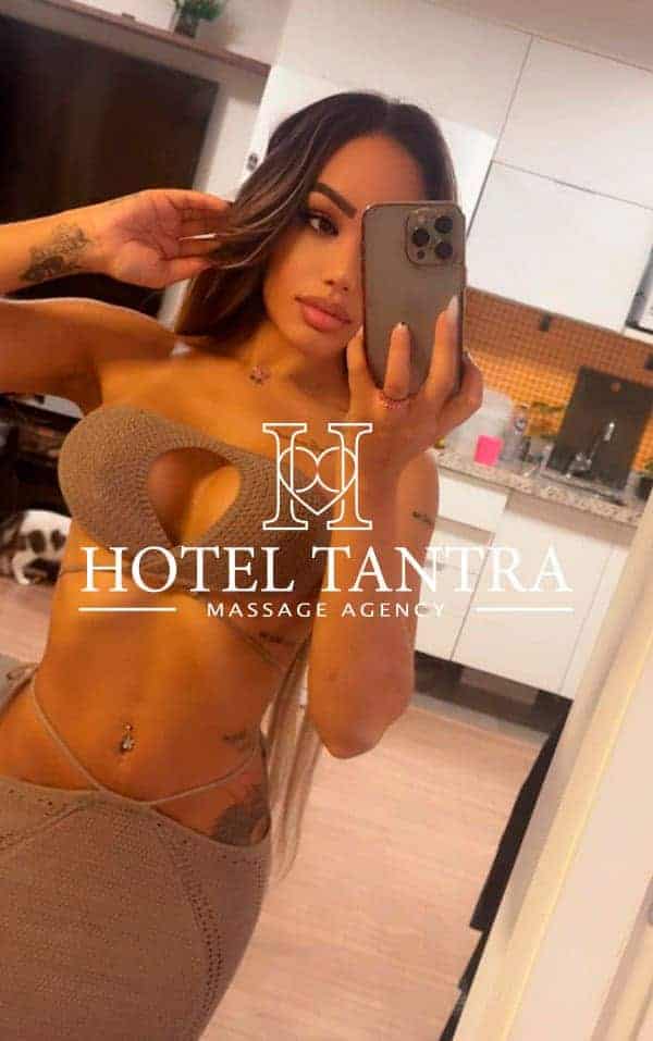 escort-madrid-marjory-tantra-outcall - Erotic Massage & Outcall Massage in Madrid Beige bikini-clad woman taking a mirror selfie in a modern kitchen, with the 'Hotel Tantra Massage Agency' logo overlaid.