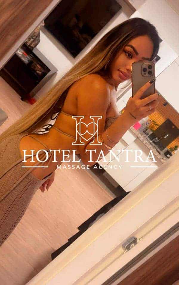 Woman in a beige bikini taking a mirror selfie in a hotel room with 'Hotel Tantra Massage Agency' logo overlay.