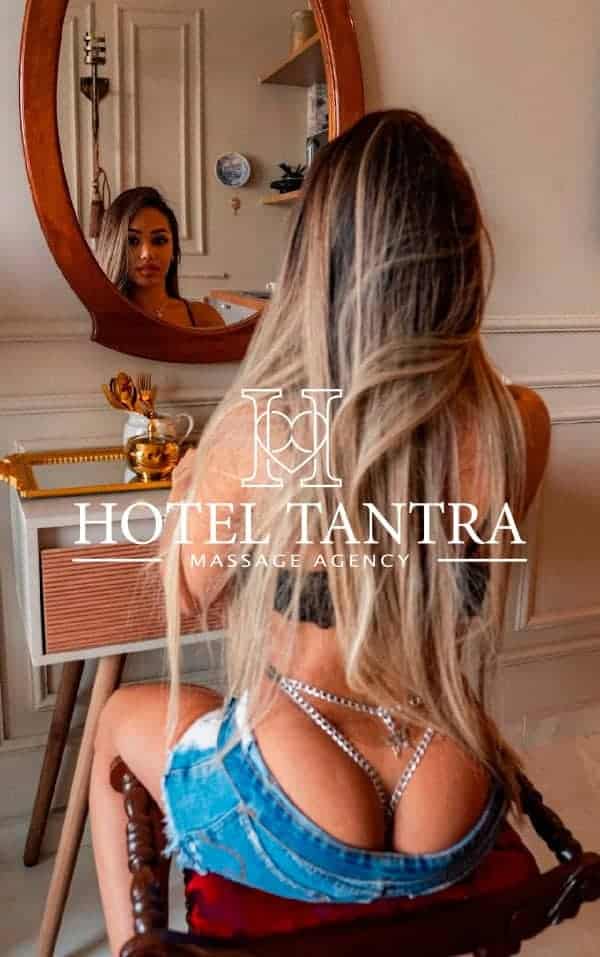 marjory-outcall-madrid-incall - Erotic Massage & Outcall Massage in Madrid Rear view of a woman with long blond hair facing a mirror in a vanity room; overlay reads 'Hotel Tantra Massage Agency'.