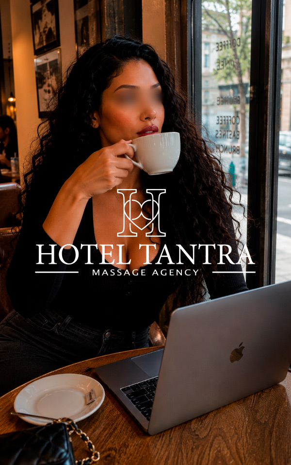 Woman with long dark hair sipping coffee at a cafe, laptop open on the table; overlay reads 'HOTEL TANTRA MASSAGE AGENCY'.