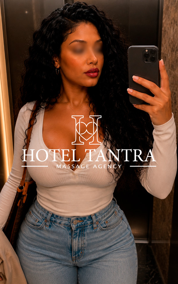 Woman taking a mirror selfie in an elevator; wearing a white ribbed top and jeans, with the overlay text 'Hotel Tantra Massage Agency'.