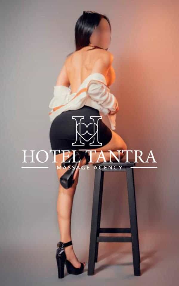 Back view of a topless woman in a short black skirt and high heels, draped in a white shirt, seated on a stool with a promotional logo reading 'Hotel Tantra Massage Agency' overlaid