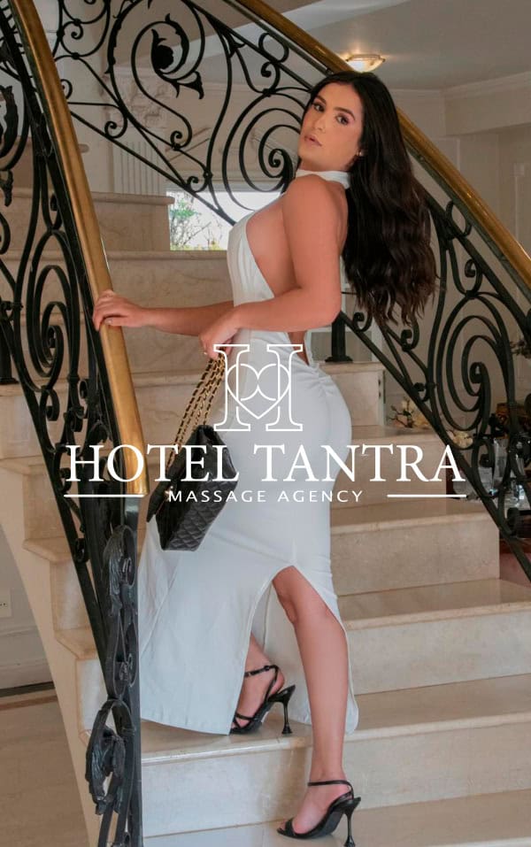 Woman in a white dress posing on a hotel staircase with a HOTEL TANTRA MASSAGE AGENCY logo overlay.