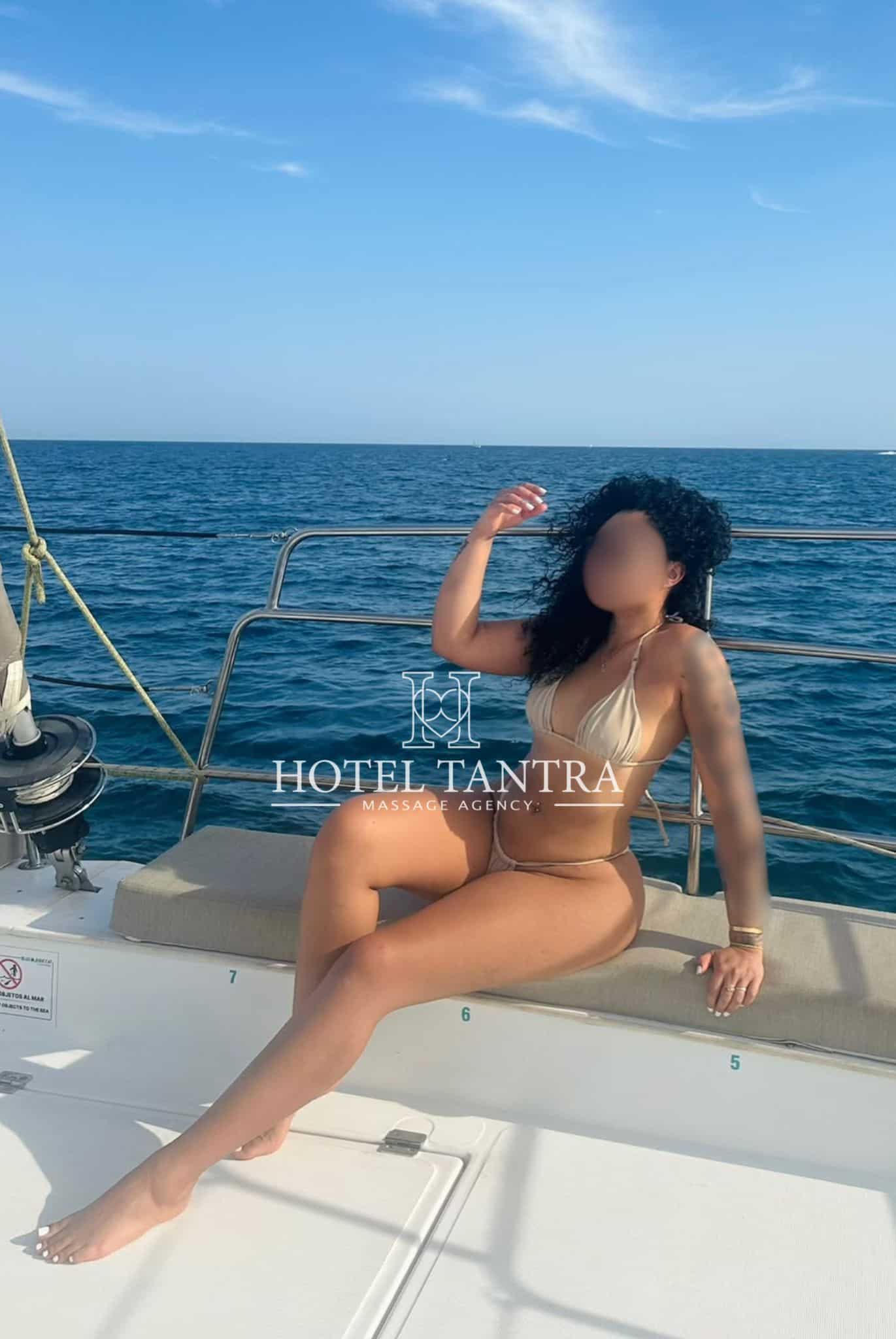 Woman in a beige bikini posing on a boat deck, with the blue ocean and sky in the background; watermark reads HOTEL TANTRA.