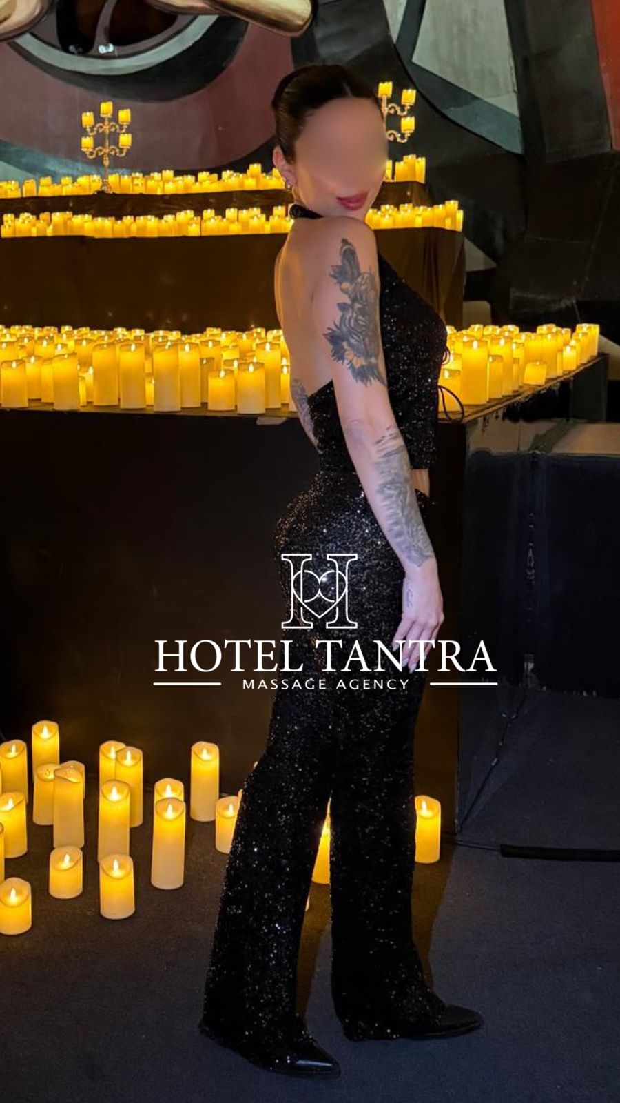Woman in a sparkly black jumpsuit stands sideways among lit candles in a dim venue, showing tattooed arm and blurred face, with 'Hotel Tantra Massage Agency' watermark.