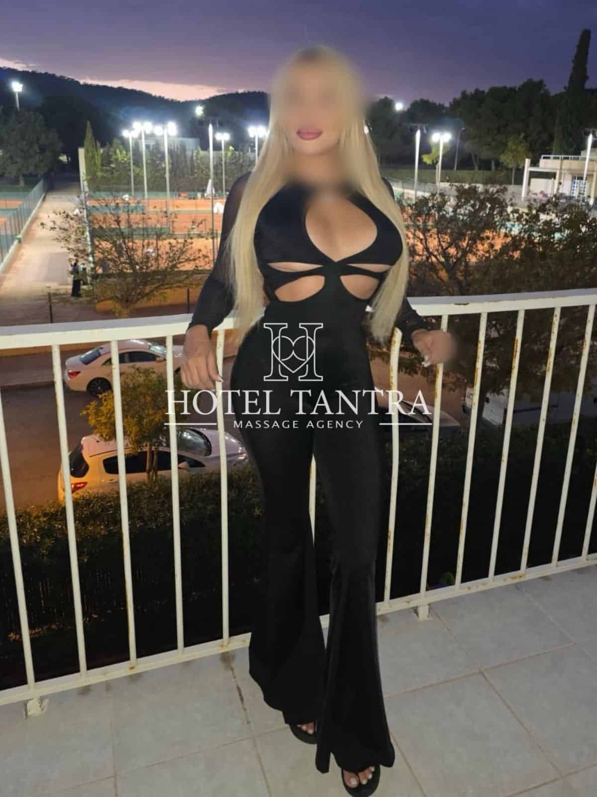 Woman in a black cut-out jumpsuit posing on a balcony at dusk, with a watermark reading 'Hotel Tantra Massage Agency' overlaid on her image