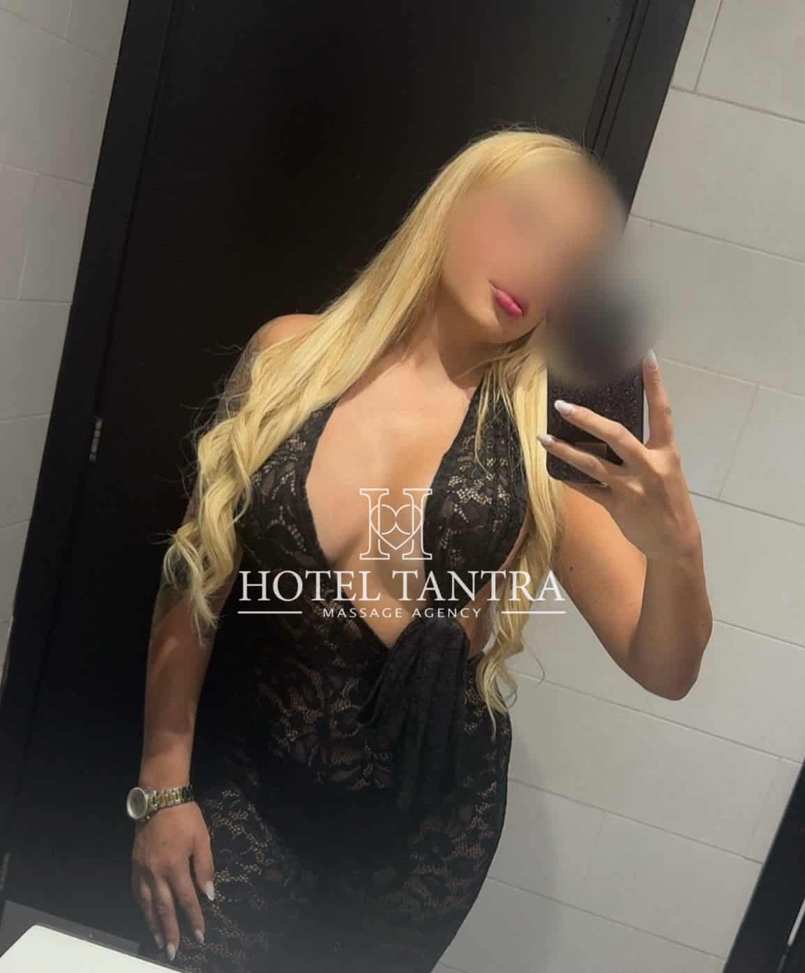 Blonde woman in a black lace dress takes a mirror selfie in a bathroom; watermark reads HOTEL TANTRA MASSAGE AGENCY across the chest area.