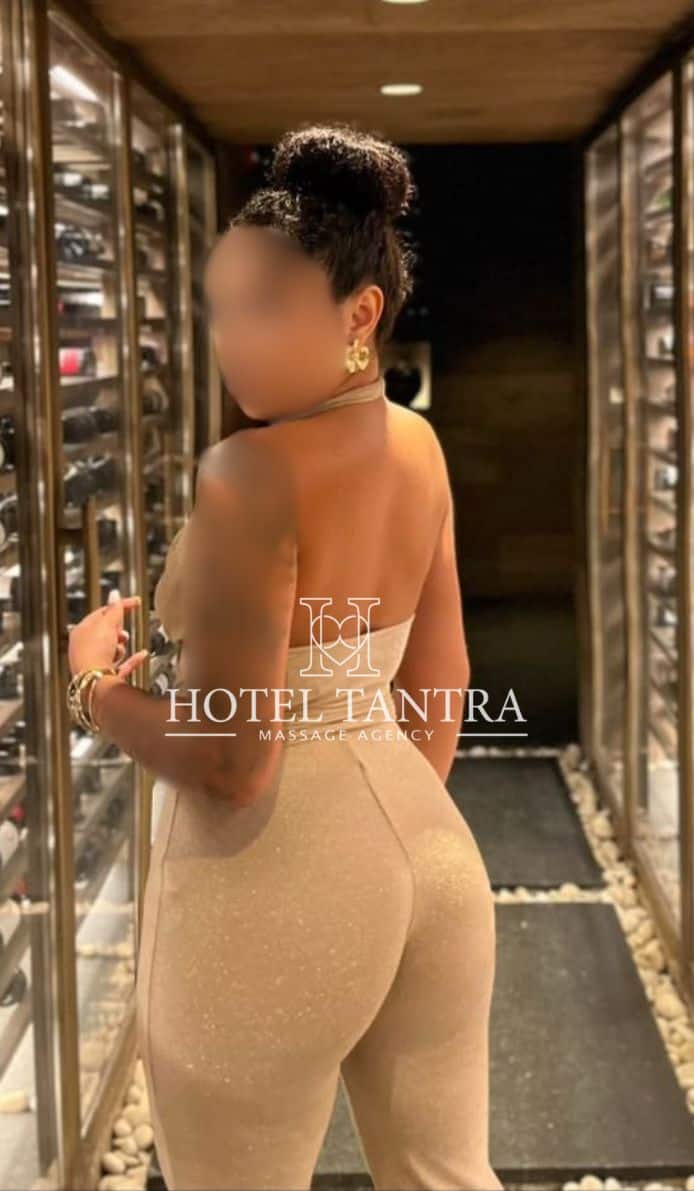 Woman with a bun, wearing a backless beige jumpsuit, standing in a wine cellar hallway.