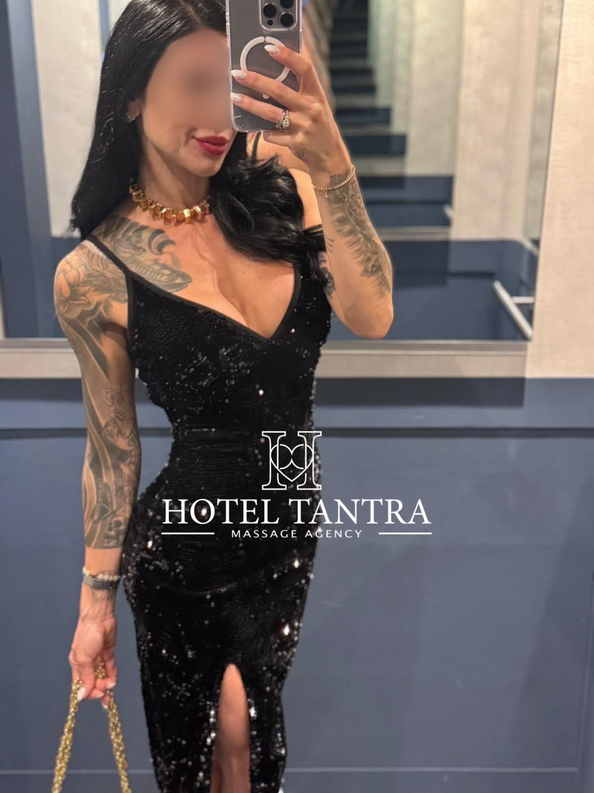 Mirror selfie of a tattooed woman in a black sequin dress, holding a smartphone with a ring grip; gold necklace and bracelets, red lipstick, watermark says 'Hotel Tantra Massage Agency'.