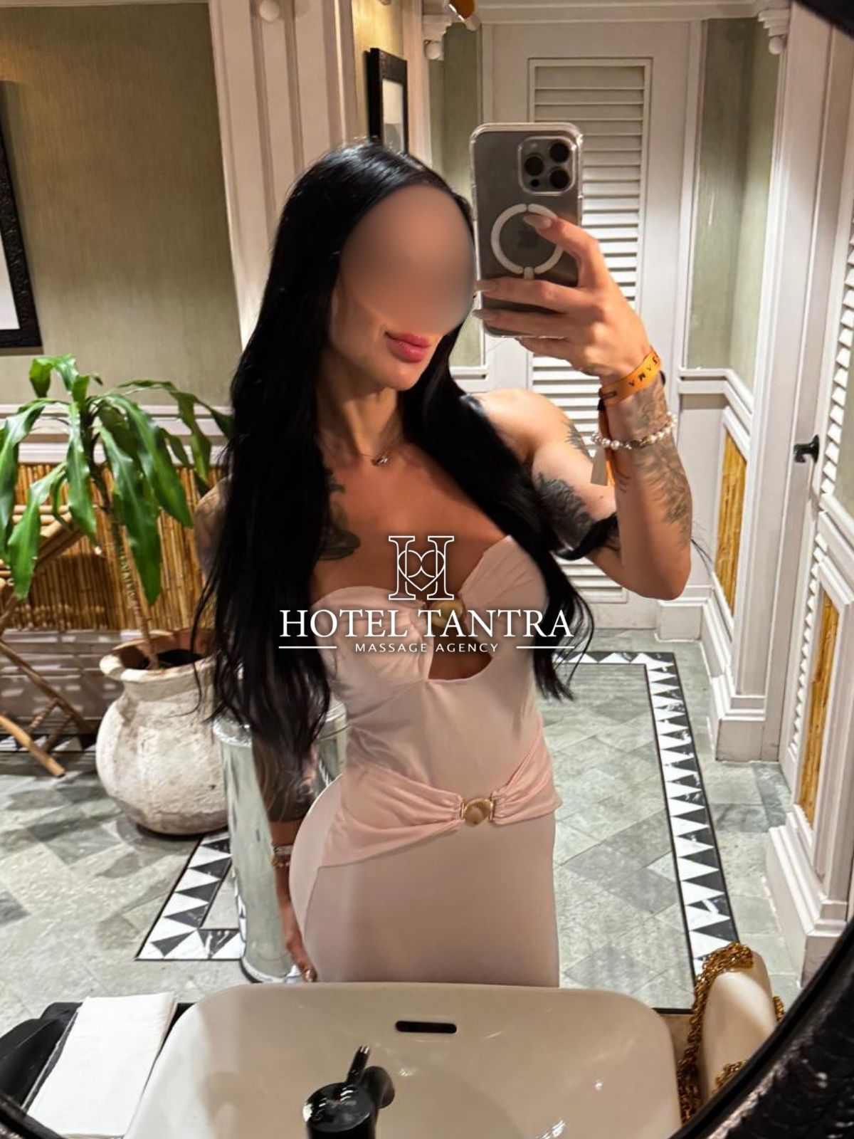 Selfie in a hotel bathroom: tattooed woman with long black hair in a pale pink strapless dress, holding a phone in front of a mirror; watermark reads Hotel Tantra Massage Agency.