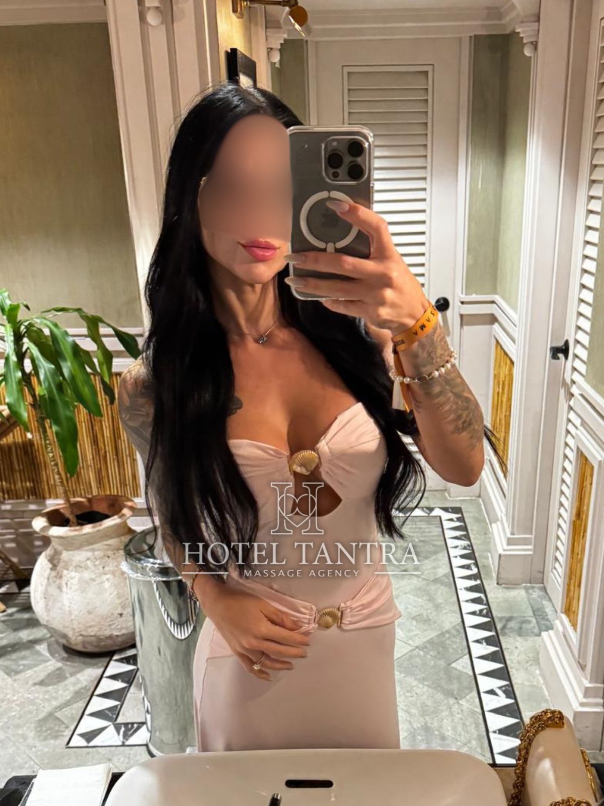 Blurred-face woman in a pale pink strapless dress taking a mirror selfie in a hotel bathroom, holding a phone with a ring grip; plants and decorative vanity in background, watermark reading 'Hotel Tantra'.