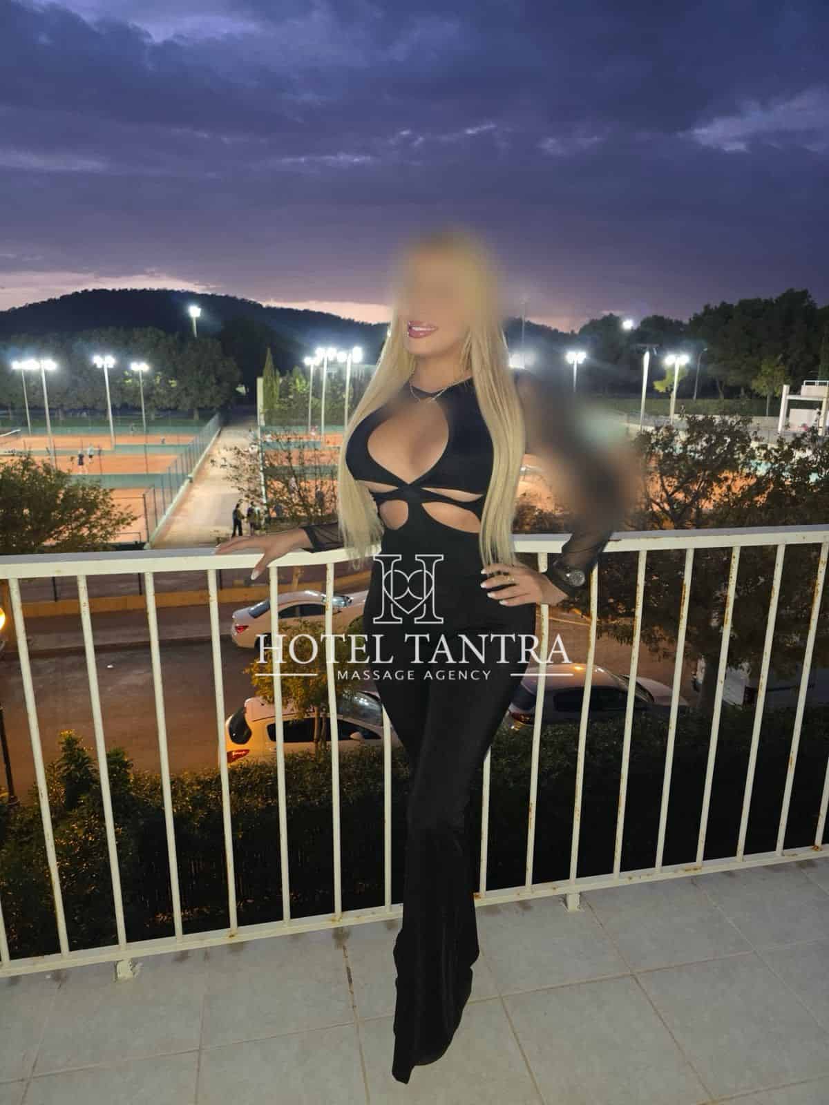 Woman in a black cut-out dress posing on a balcony at dusk, with tennis courts and lights in the background (watermark: HOTEL TANTRA).