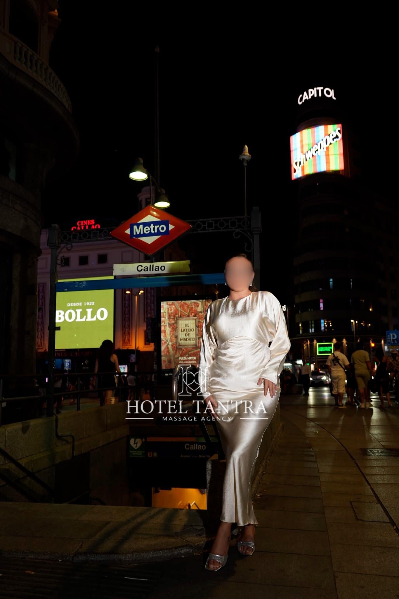 Woman in a white satin dress posing on a lit city sidewalk at night, with a Metro Callao sign and tall illuminated signs in the background.