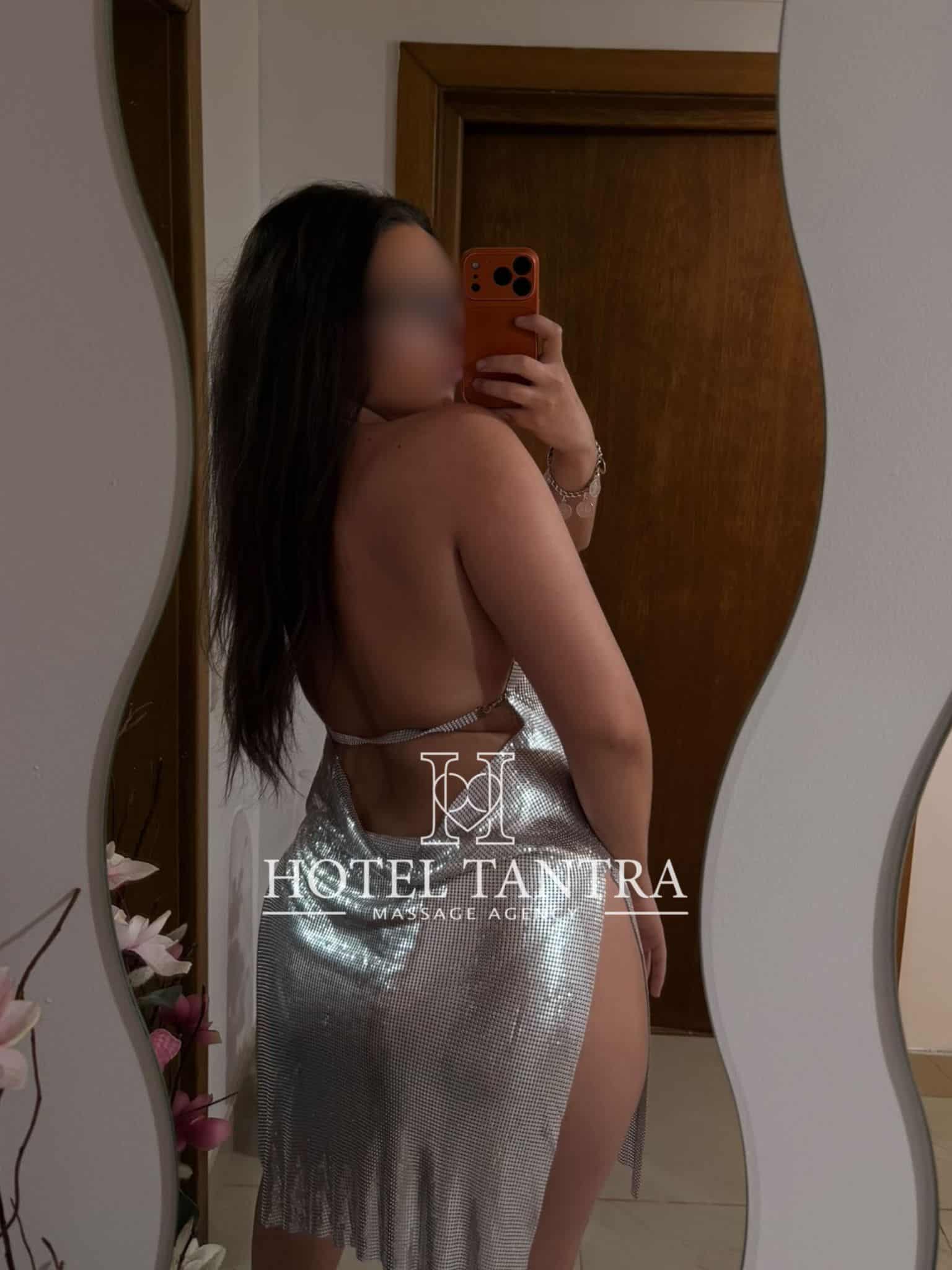 Woman with long dark hair stands topless from behind in a doorway mirror, holding an orange phone to take a selfie; silver metallic dress drapes over her hips; watermark reads 'HOTEL TANTRA MASSAGE'.
