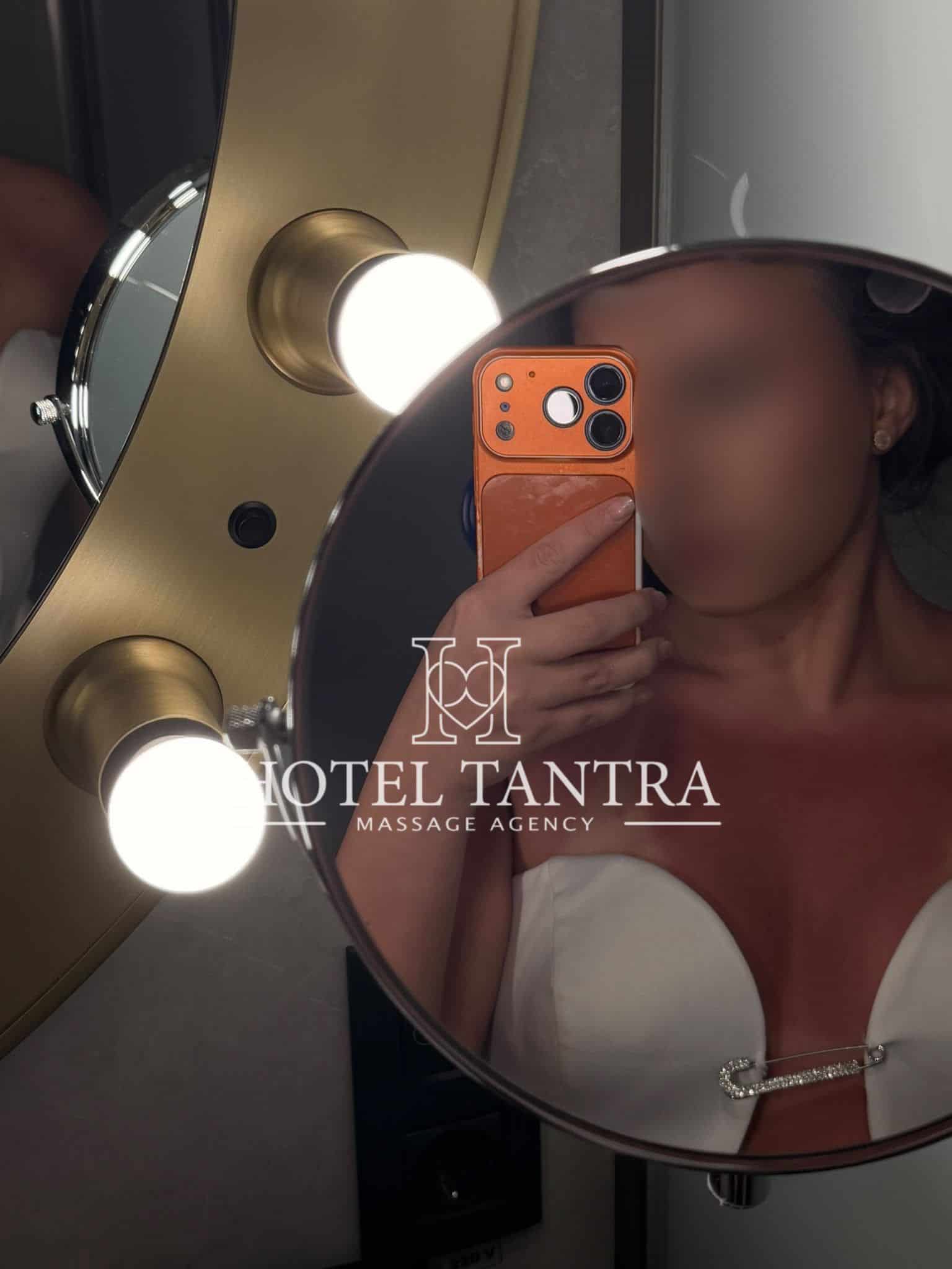 Person takes a mirror selfie in a bathroom, holding an orange iPhone, with round vanity lights around a gold-toned mirror and a watermark reading HOTEL TANTRA.