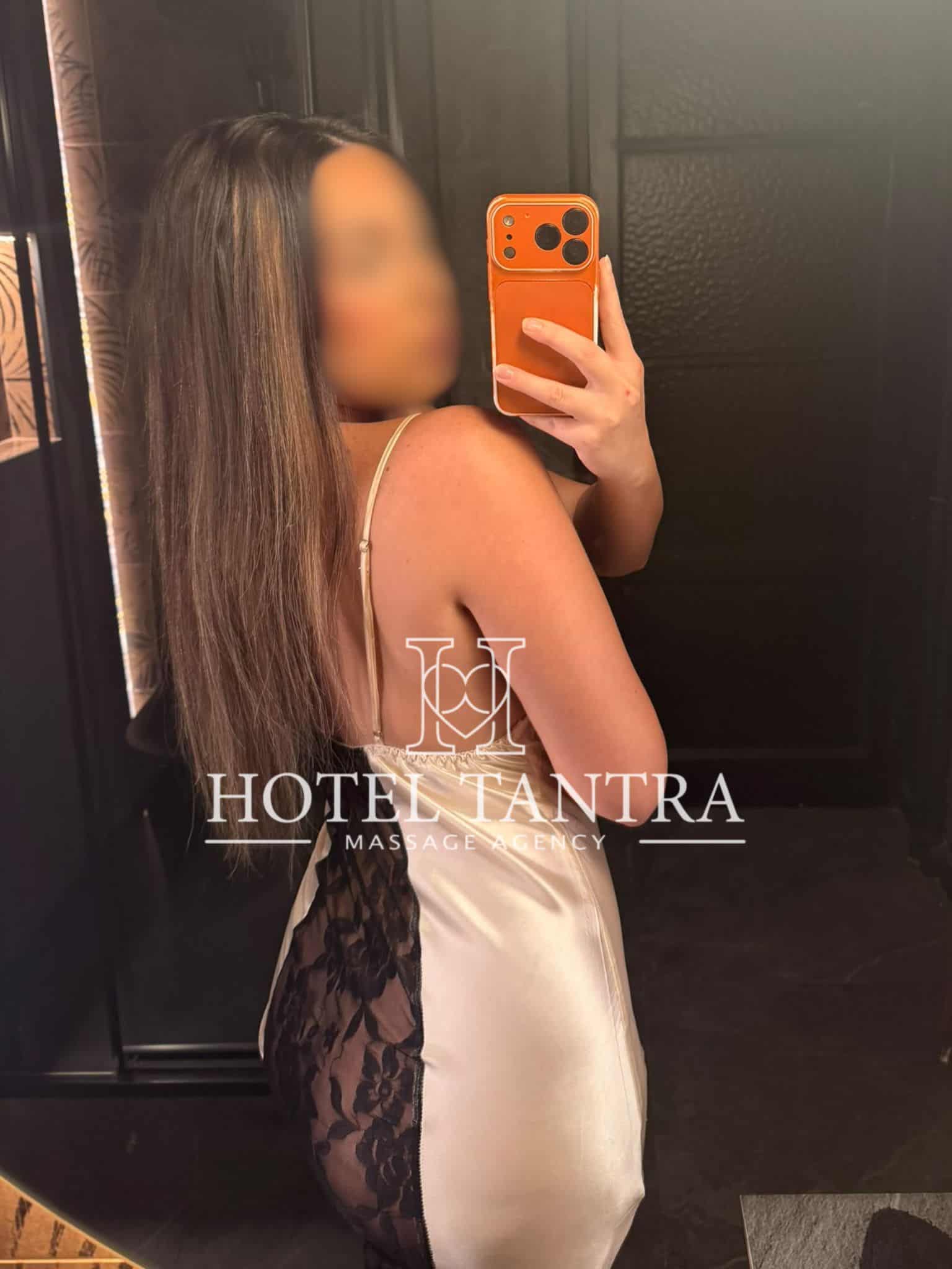 Woman in a beige satin dress with black lace taking a mirror selfie, holding an orange phone case in a dim, reflective surface with 'Hotel Tantra' branding at the bottom.