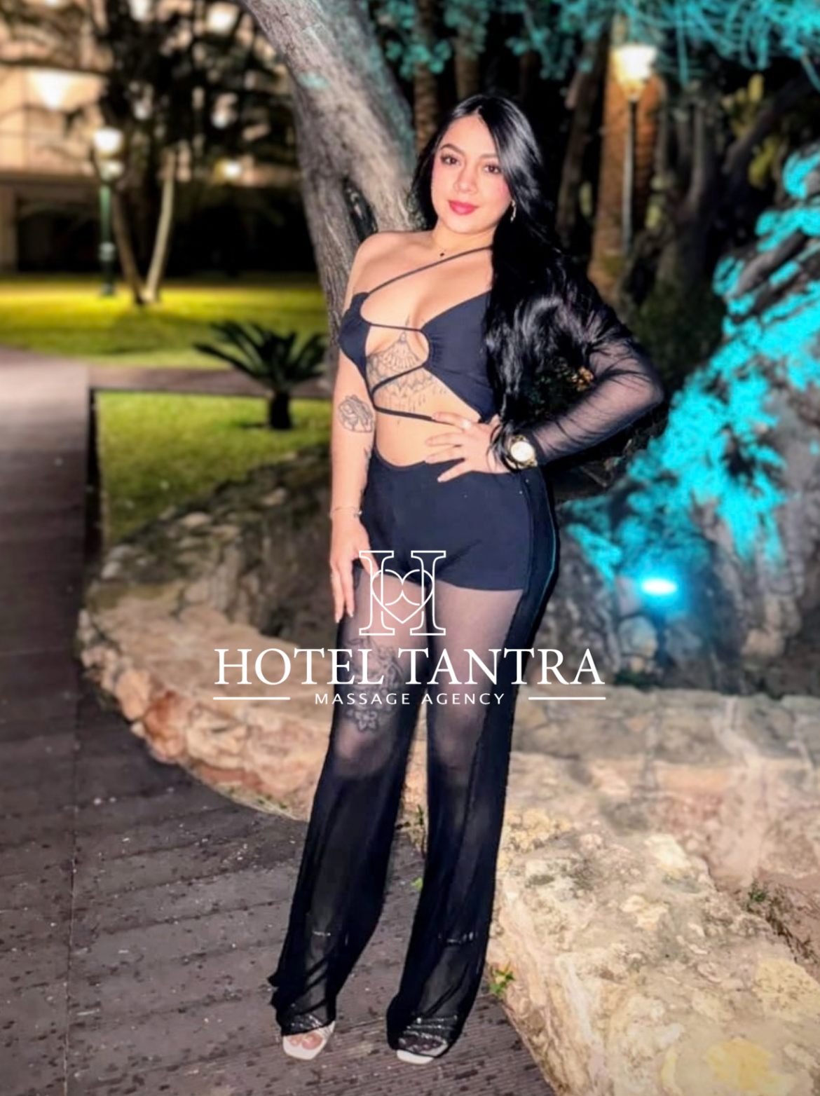 Woman in a black cut-out outfit posing beside a tree at night, with a hotel watermark reading 'Hotel Tantra' on her legs.