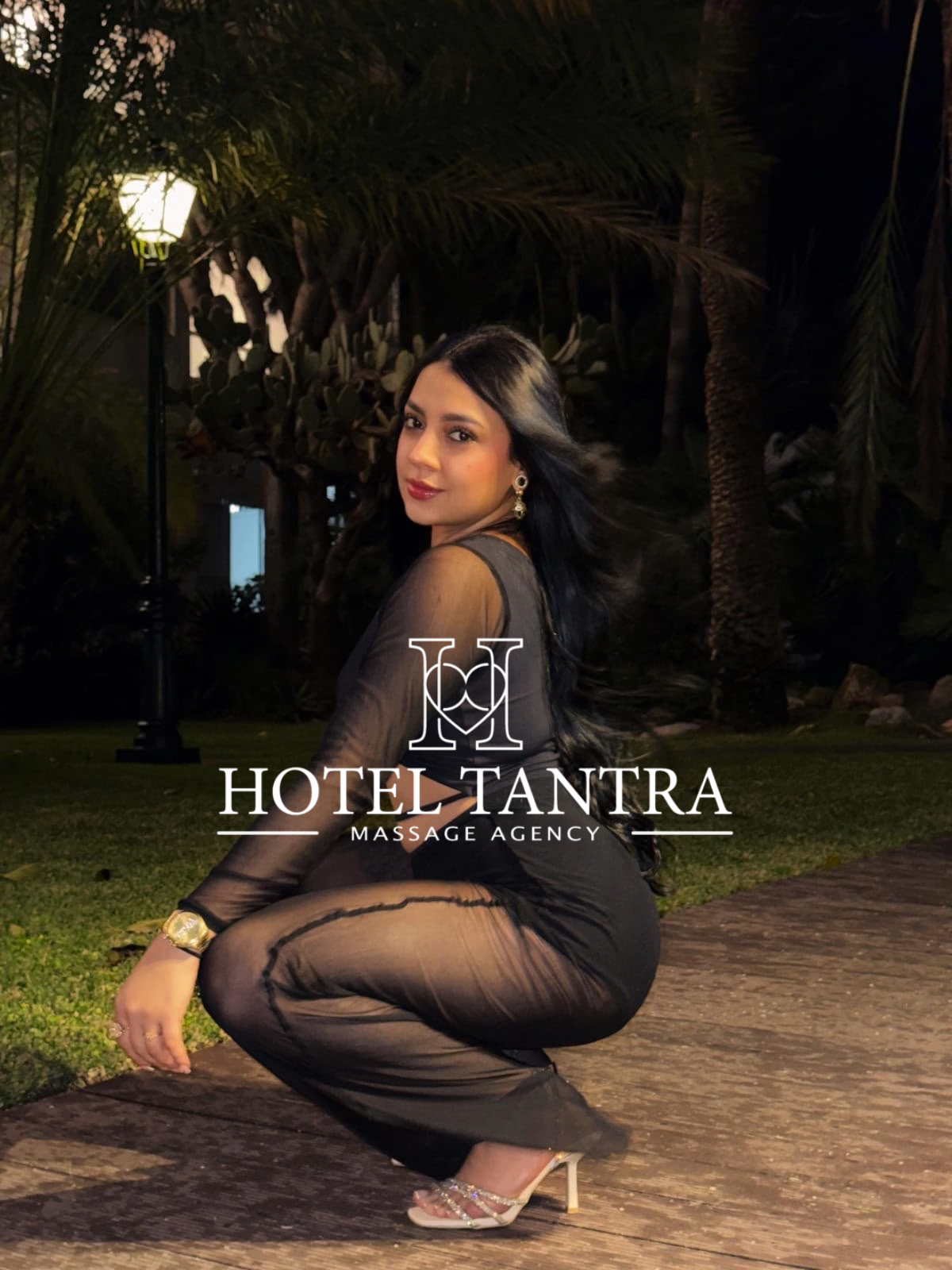 Outdoor nighttime portrait of a woman in a sheer black outfit, crouching on a path with a Hotel Tantra Massage Agency logo overlaid