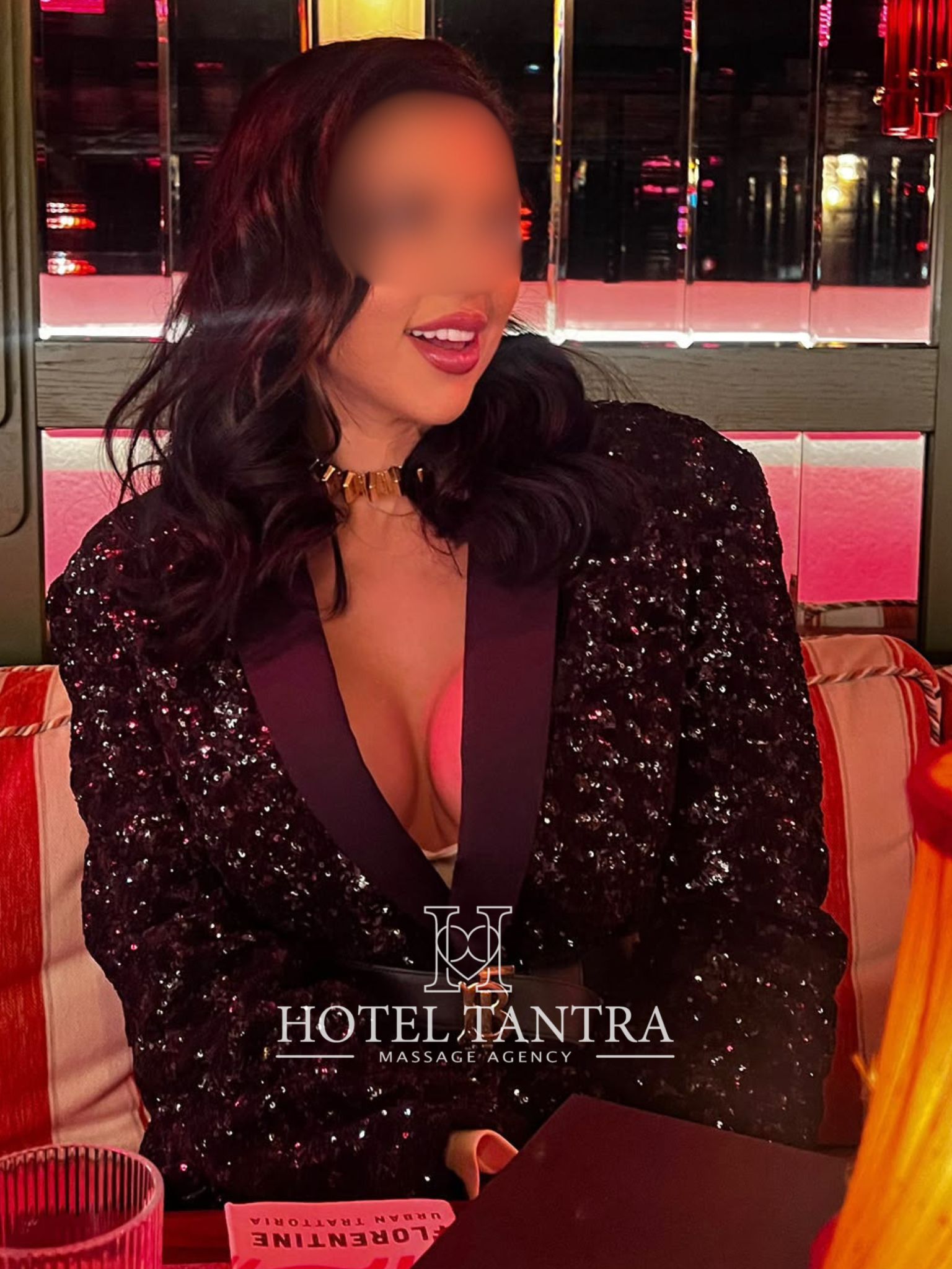 Woman in a black sequined blazer with a gold choker sits at a table in a red-lit lounge; face blurred for privacy.