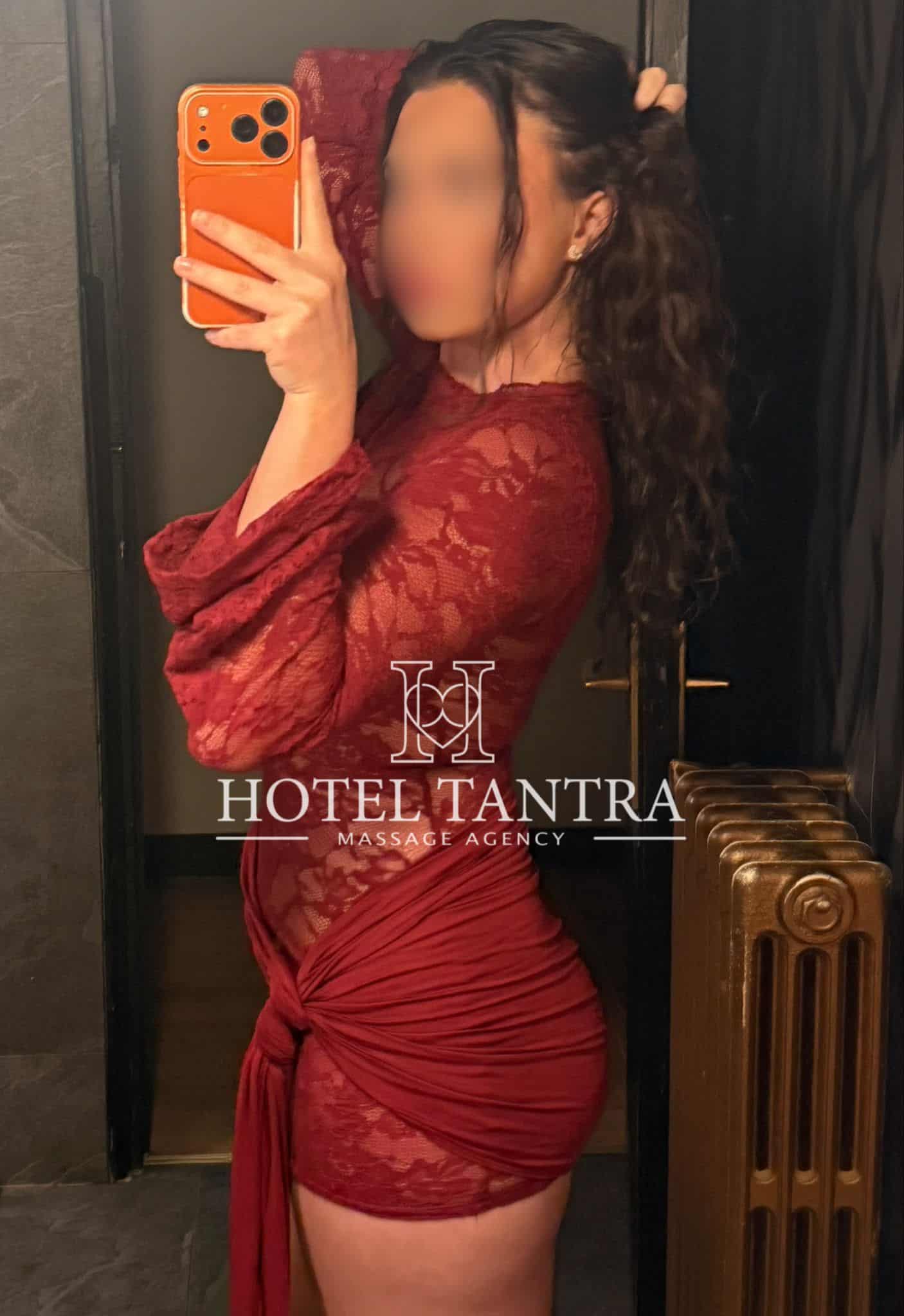 Person in a red lace dress taking a mirror selfie in an elevator, holding an orange smartphone; watermark reads 'Hotel Tantra Massage Agency'.