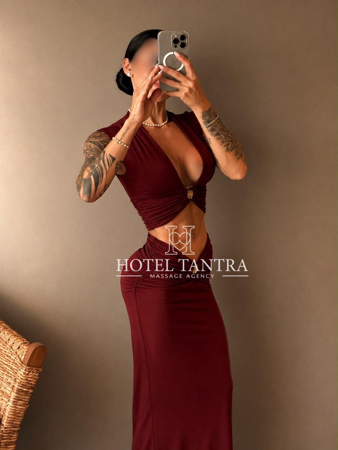 Woman in a burgundy cut‑out dress takes a mirror selfie; visible tattoos on both arms and a pearl necklace, with a Hotel Tantra watermark.
