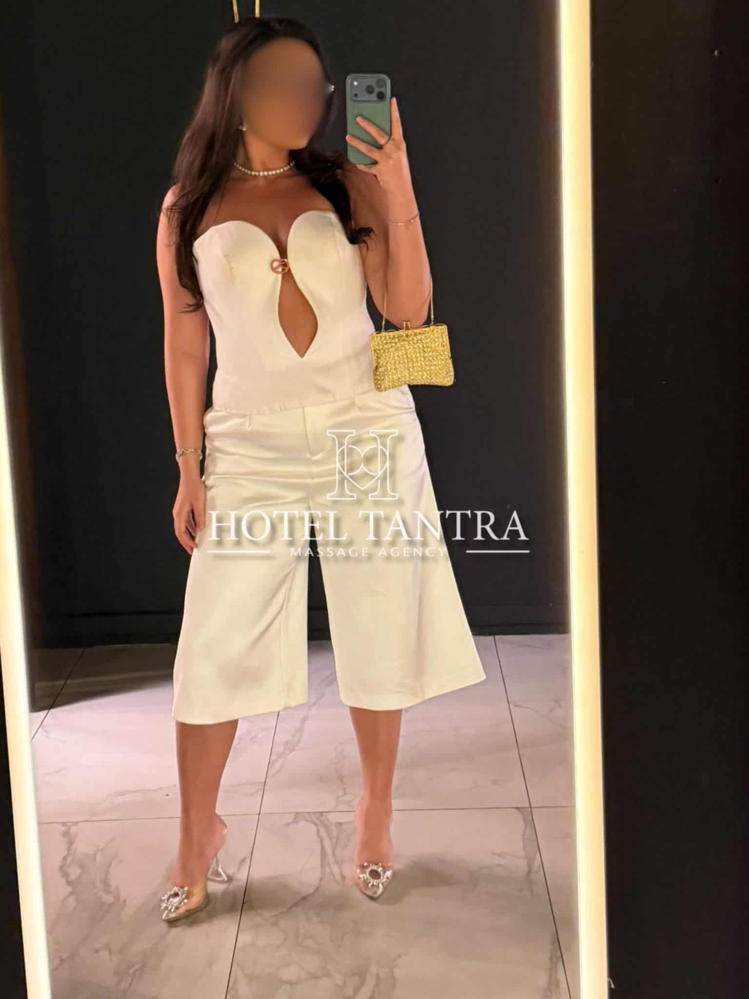 Woman in a white strapless jumpsuit with a front cutout, pearl necklace, and gold beaded clutch posing for a mirror selfie in a chic lobby.