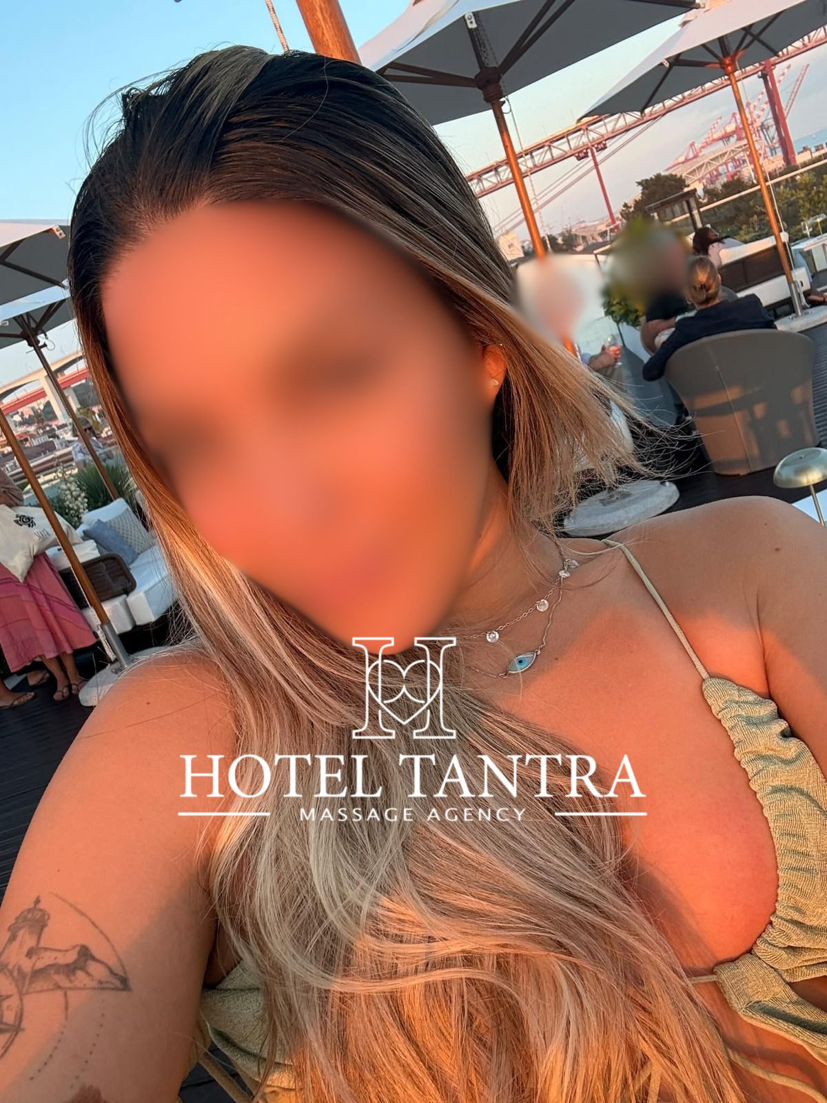 Close-up selfie of a woman with long blond hair wearing a bikini top at an outdoor hotel lounge with large umbrellas and guests in the background; 'Hotel Tantra' watermark visible.